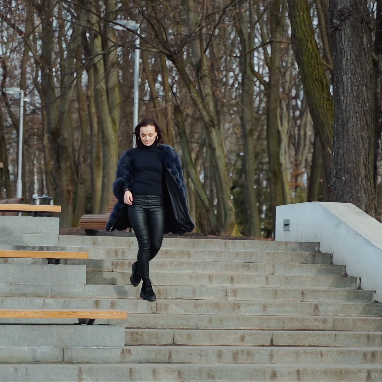 Beautiful girl in the park. Happy young woman goes down the stairs in the park. Attractive model in fur coat. Slow motion.