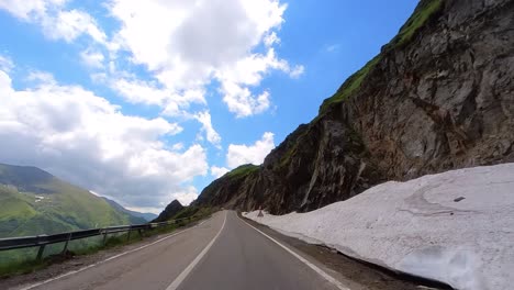 A-Slow-Drive-Up-the-Transfagarasan-Highway,-Driver’s-Point-of-View,-Surrounded-by-Tall-Mountain-Sides-with-Patches-of-Snow-and-a-Clear-Blue-Sky-in-Romania