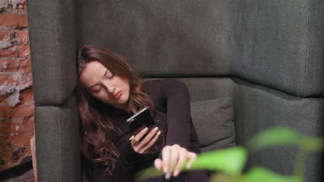 young woman in black outfit relaxes on green cushioned seat, holding smartphone with one hand and playing with fingers mid-air, slightly leaning to side near table. cozy indoor space with calm mood