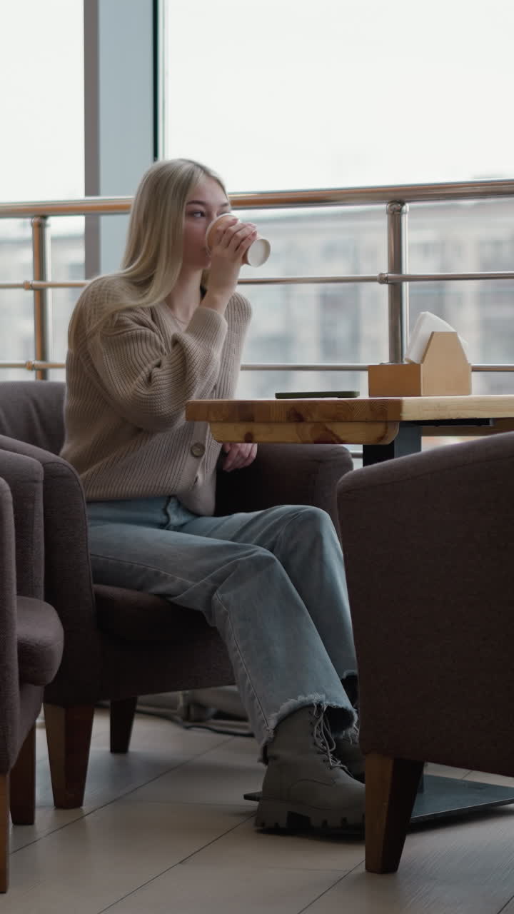joven bebiendo té de su taza, dejándola caer en la mesa mientras mira hacia afuera a través de vidrio transparente en el centro comercial, con una chaqueta de invierno colgada en la silla