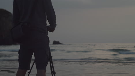 Hombre-Sosteniendo-Una-Cámara-En-Un-Trípode-En-La-Playa-Durante-La-Puesta-De-Sol-En-Cornualles,-Reino-Unido---Plano-Medio