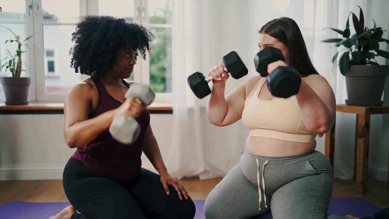 Sorrindo, duas mulheres ajoelhadas em tapetes roxos, fazendo rosca com halteres em roupa de ginástica e incentivando uma à outra.