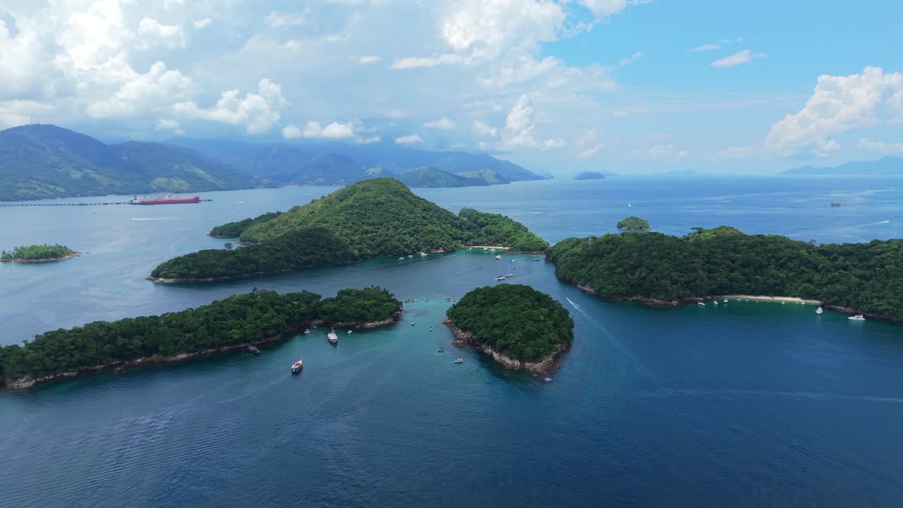 เกาะเล็กๆ ที่มีพืชพรรณปกคลุมในอิลย่ากรังดี, ลากัวอาซุล, อังกร้าโดสเรส์, ริโอเดจาเนโร ภาพมุมกว้างทางอากาศที่แพนออกเผยให้เห็นทิวทัศน์ของหมู่เกาะที่ล้อมรอบด้วยมหาสมุทรสีครามและเรือในวันที่เมฆเป็นบางส่วน