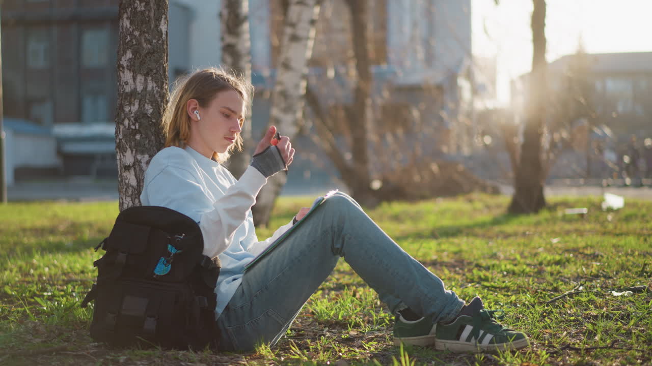 어린 학생이 나무 근처 야외에서 스케치해요. 편안하게 음악과 자연을 즐기며 그림을 그리는 십 대, 부드러운 햇살 아래 자작나무 옆에서 클립보드에 스케치하는 편안한 청소년