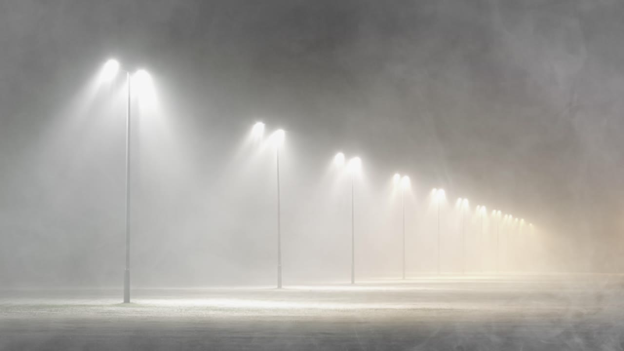 A row of streetlights vanishes into the distance, their beams cutting through thick, rolling fog on an empty street for a mysterious, ethereal mood
