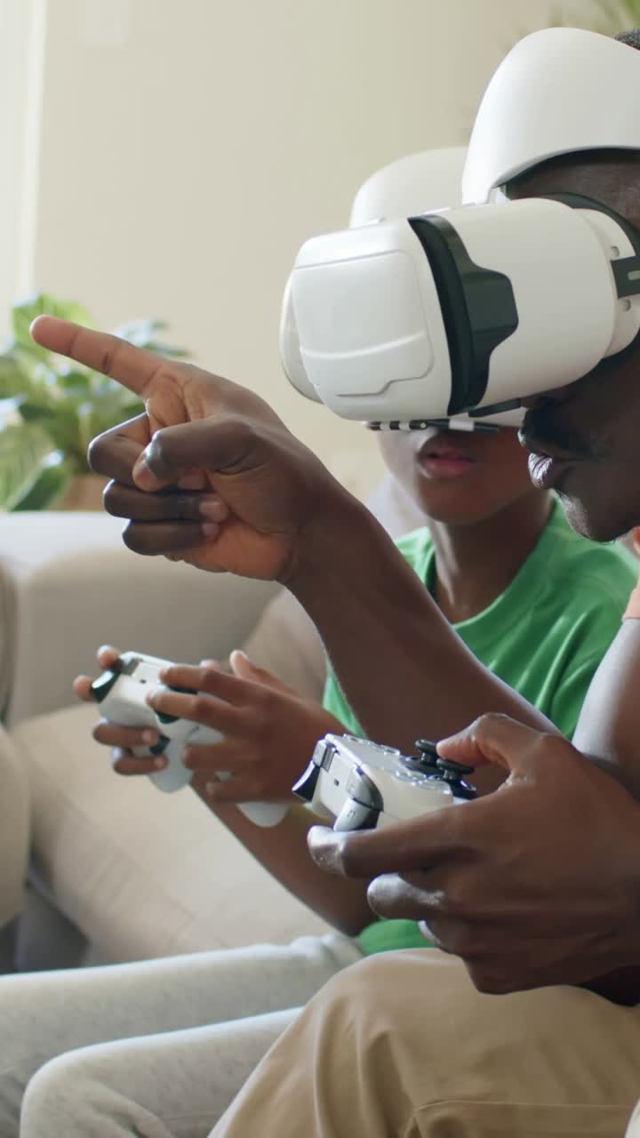 Video verticale: Gioco che inizia, padre e figlio che indicano e premono i controller sul divano, con maglietta arancione VR, per divertimento.