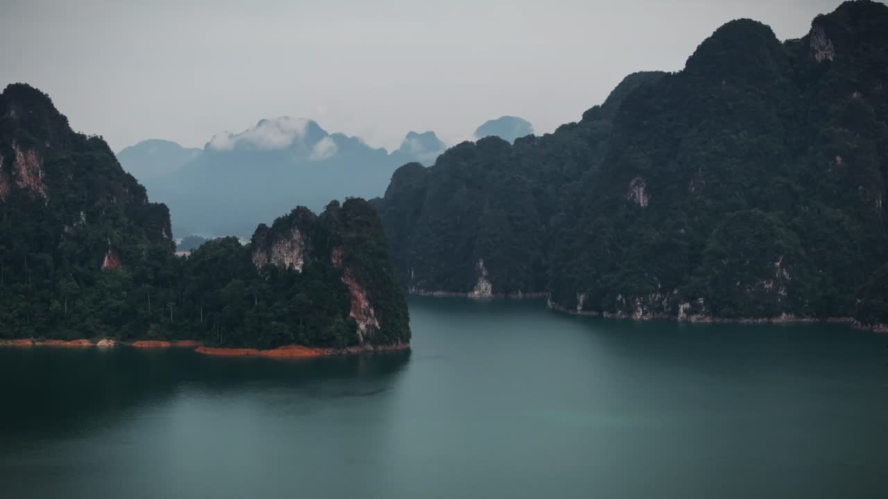 Cinematic drone footage of Khao Sok National Park in Thailand, showcasing dramatic limestone cliffs and calm teal waters under a cloudy sky