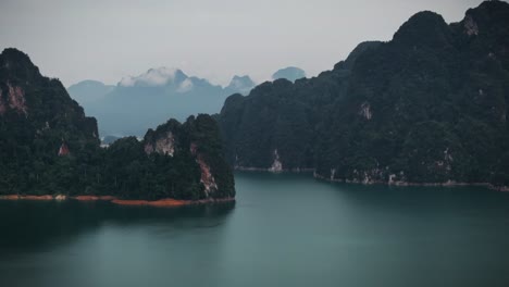 cinematic drone footage of khao sok national park in thailand, showcasing dramatic limestone cliffs and calm teal waters under a cloudy sky