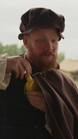 hombre barbudo con ropa medieval y gorra abre una botella de poción mágica y comienza a beber. el escudero utiliza medios mágicos para mantener una buena forma física en el pueblo