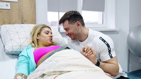 caring husband talks and smiles to his pregnant wife lying in bed. couple expecting the newborn in the maternity hospital ward.