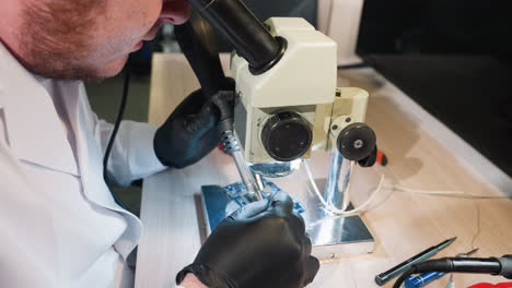 close-up view of a technician in a white lab coat using a microscope and soldering tools to inspect and repair a circuit board