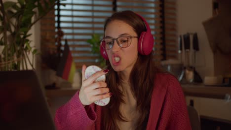 Close-up,-a-young-confident-brunette-girl-with-glasses-in-red-wireless-headphones-and-a-sweater-studies-a-foreign-language-German-and-looks-at-herself-with-the-help-of-a-mirror-while-pronouncing-words-in-a-modern-apartment-in-the-kitchen