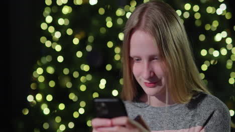 una adolescente usa un teléfono inteligente en el fondo de un árbol de navidad