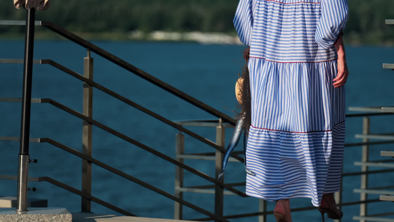 Woman in a blue gingham dress holding wide hat walking on a scenic terrace with a view of a calm body of water