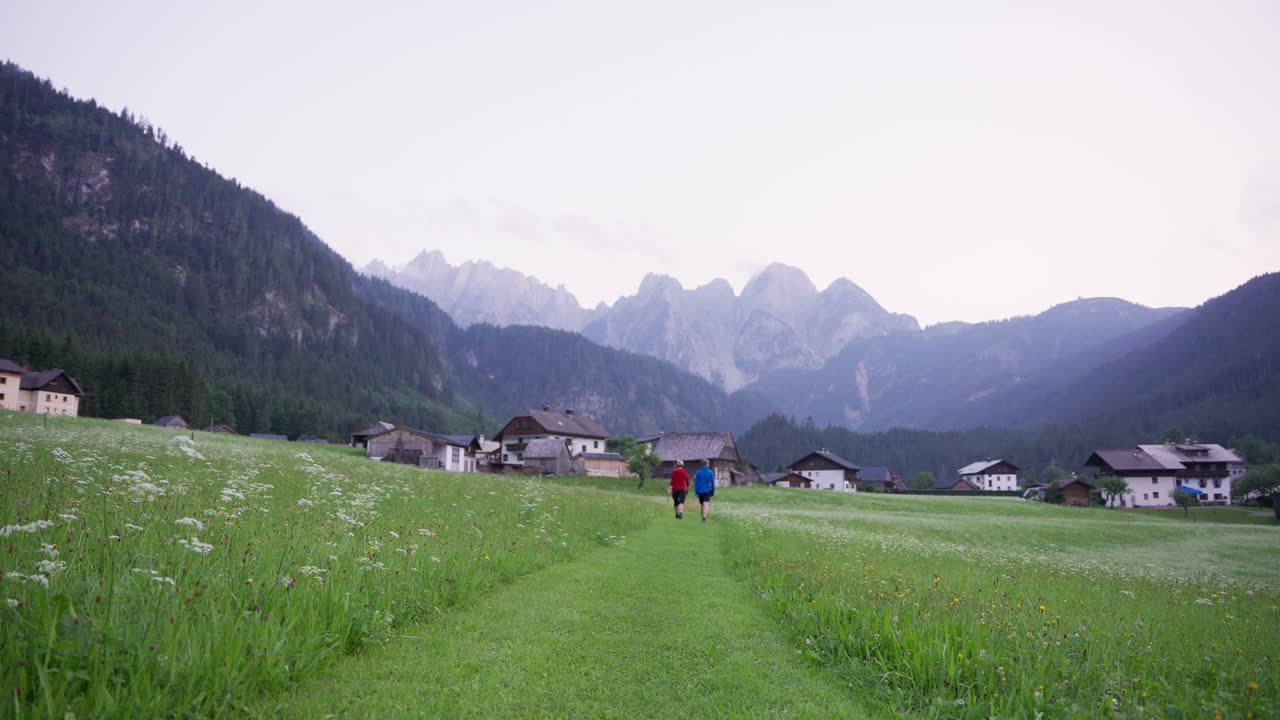 Two hikers walking towards a village with mountains in the background at sunset