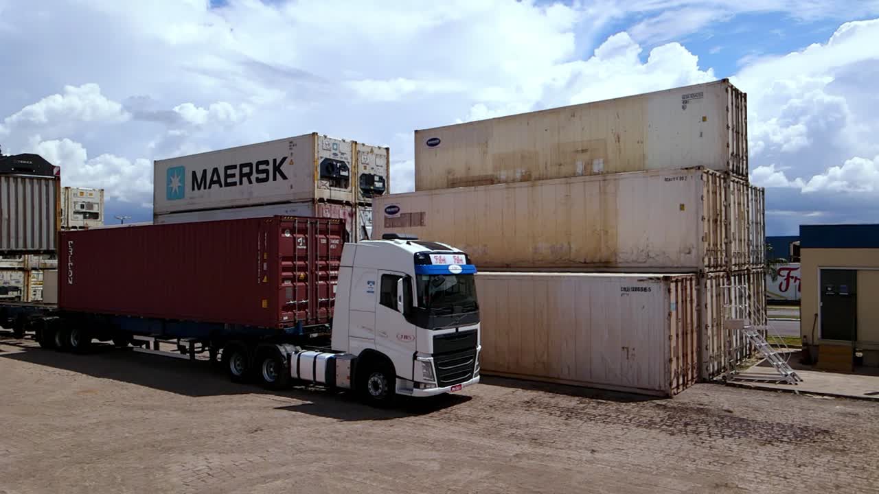 Crane places shipping container on truck in front of containers, aerial pan