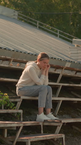 young woman sitting thoughtfully on rustic stadium bleachers, resting her chin on hands, wearing casual hoodie and jeans, she appears deep in thought, surrounded by greenery