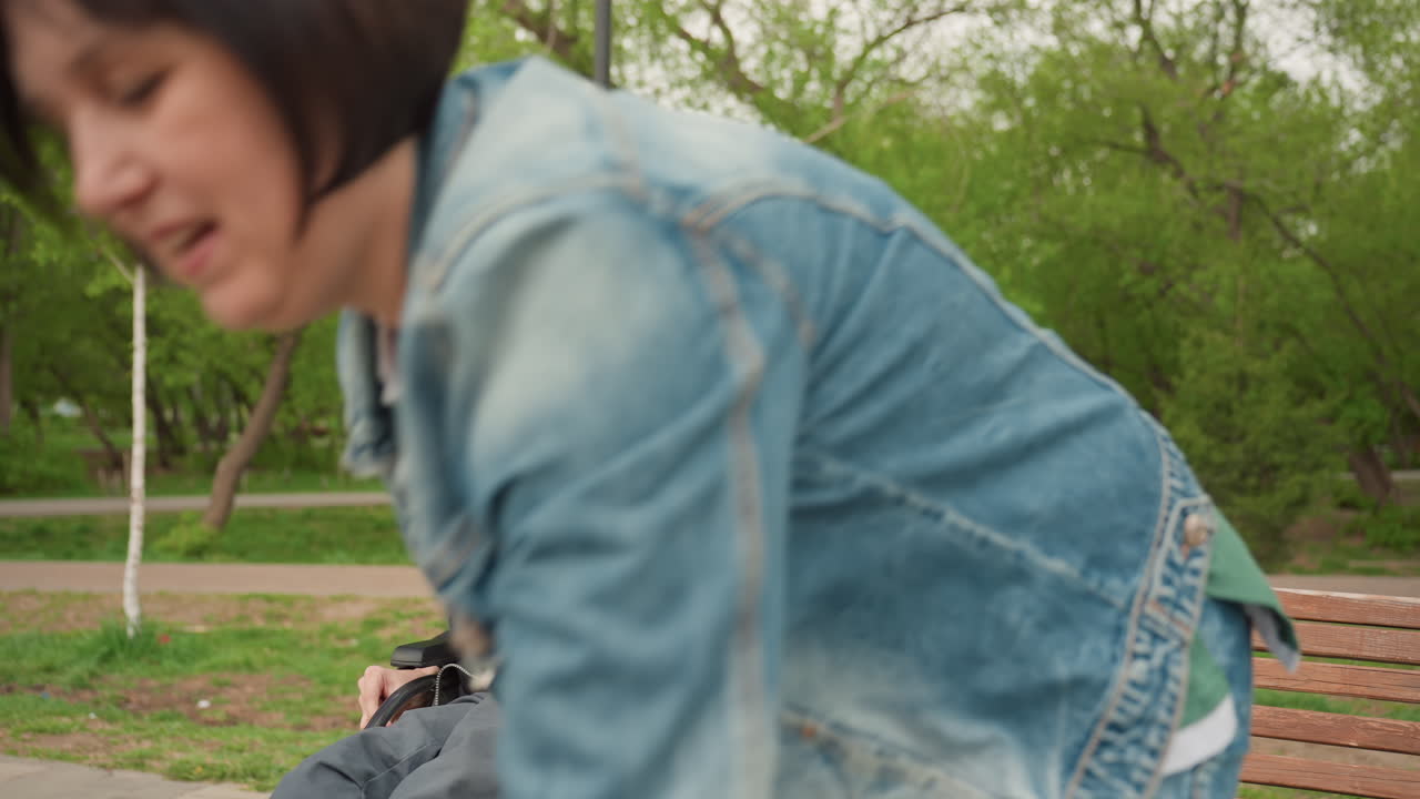 Caregiver Demonstrating Expressive Hand Gestures While Young Man In Wheelchair Watches And Copies On Bench In Park, Animated Teaching Moment, Open Palms, Engaged Faces, Practical Communication