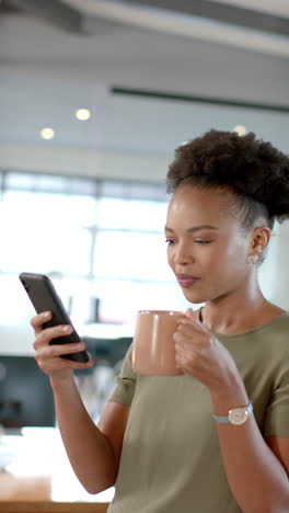 vertical video: holding smartphone and drinking coffee, woman checking messages in office