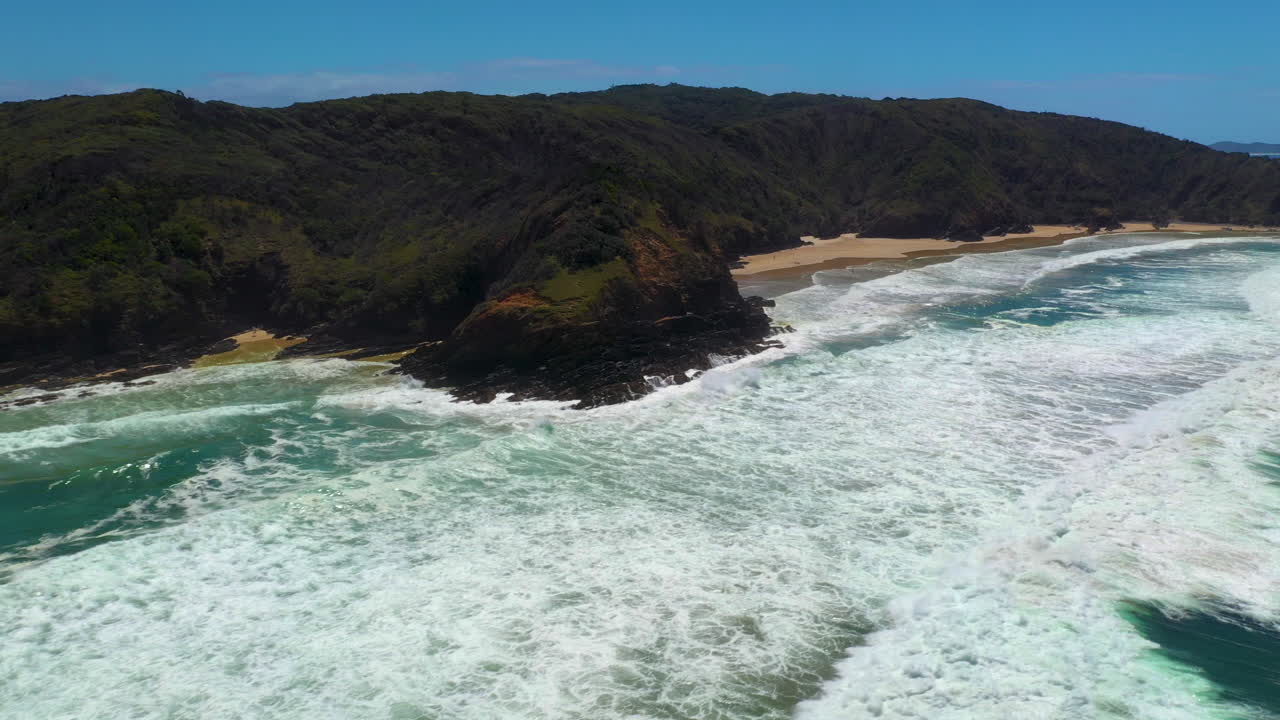Wide cinematic drone shot of crashing waves and island at Broken Head beach in Australia