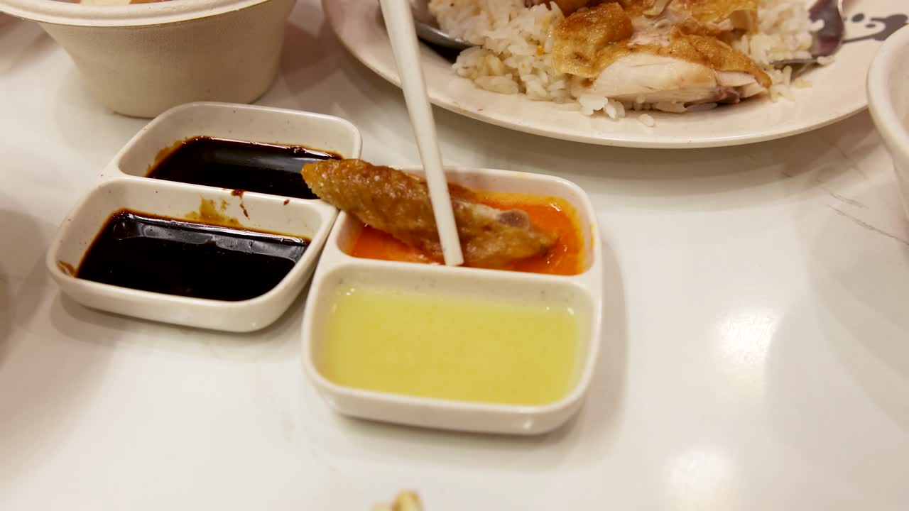 Hand uses chopsticks to dip roasted chicken into chili and ginger sauces beside Hainanese chicken rice under bright indoor lighting, static close-up view