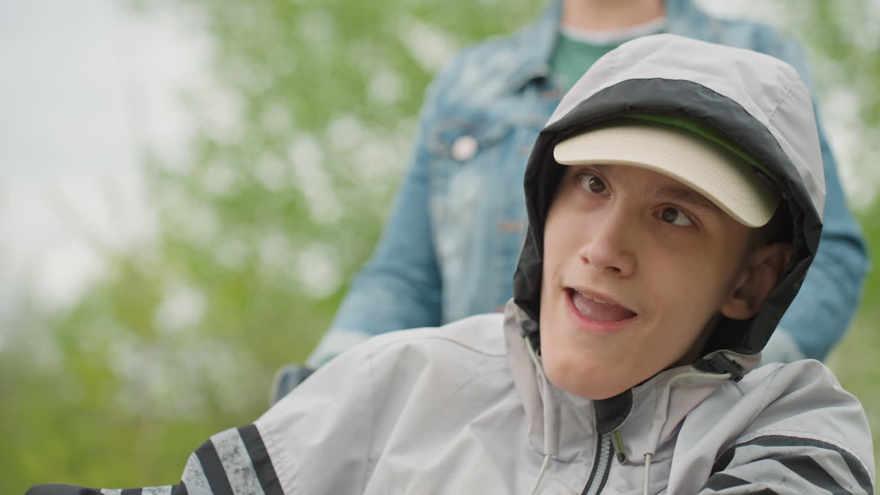 White Teen With Caregiver In Park, Denim Attendant Standing Behind Wheelchair, Attentive Listening And Gentle Interaction, Hooded Jacket And Cap, Natural Green Backdrop, Inclusive Outdoor Support