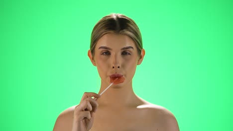 fotografía estática en cámara lenta de una joven rubia lamendo una piruleta en forma de corazón y dejando que el sabor al azúcar se derrita en sus labios frente al fondo verde