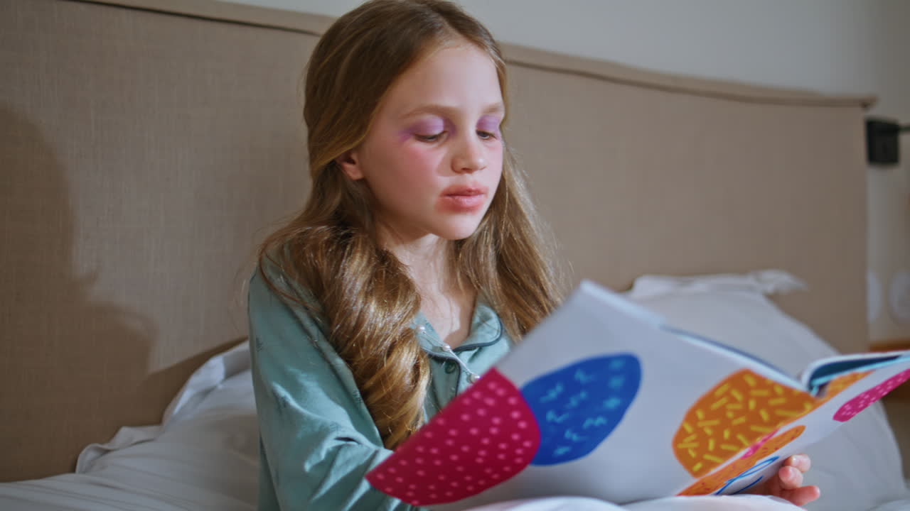 Bambino carino che sfoglia un libro e si mette un dito in bocca in primo piano in camera da letto. Bambino divertente che legge