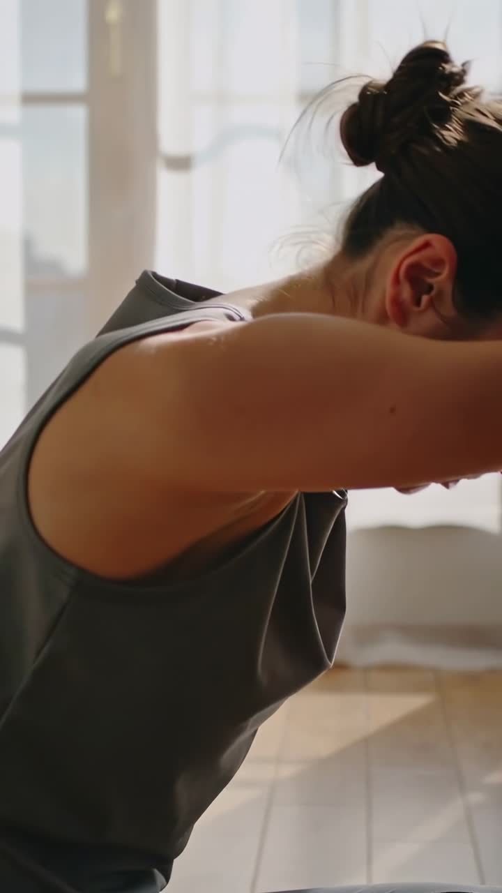 Vídeo vertical: Mulher inspirando e levantando os braços, dobrando-se para a frente em uma pose de yoga para acalmar-se perto da janela