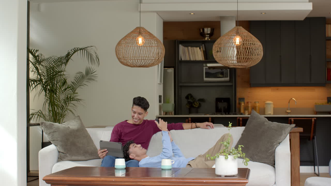 Relaxing on couch, multiracial gay couple enjoying tablet in cozy living room, at home