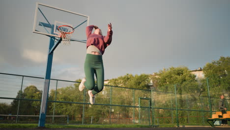 lady in outdoor court jumps, throws volleyball up, waits for it to land, then catches it, basketball hoop visible in background with people seated, greenery, and buildings surrounding the area