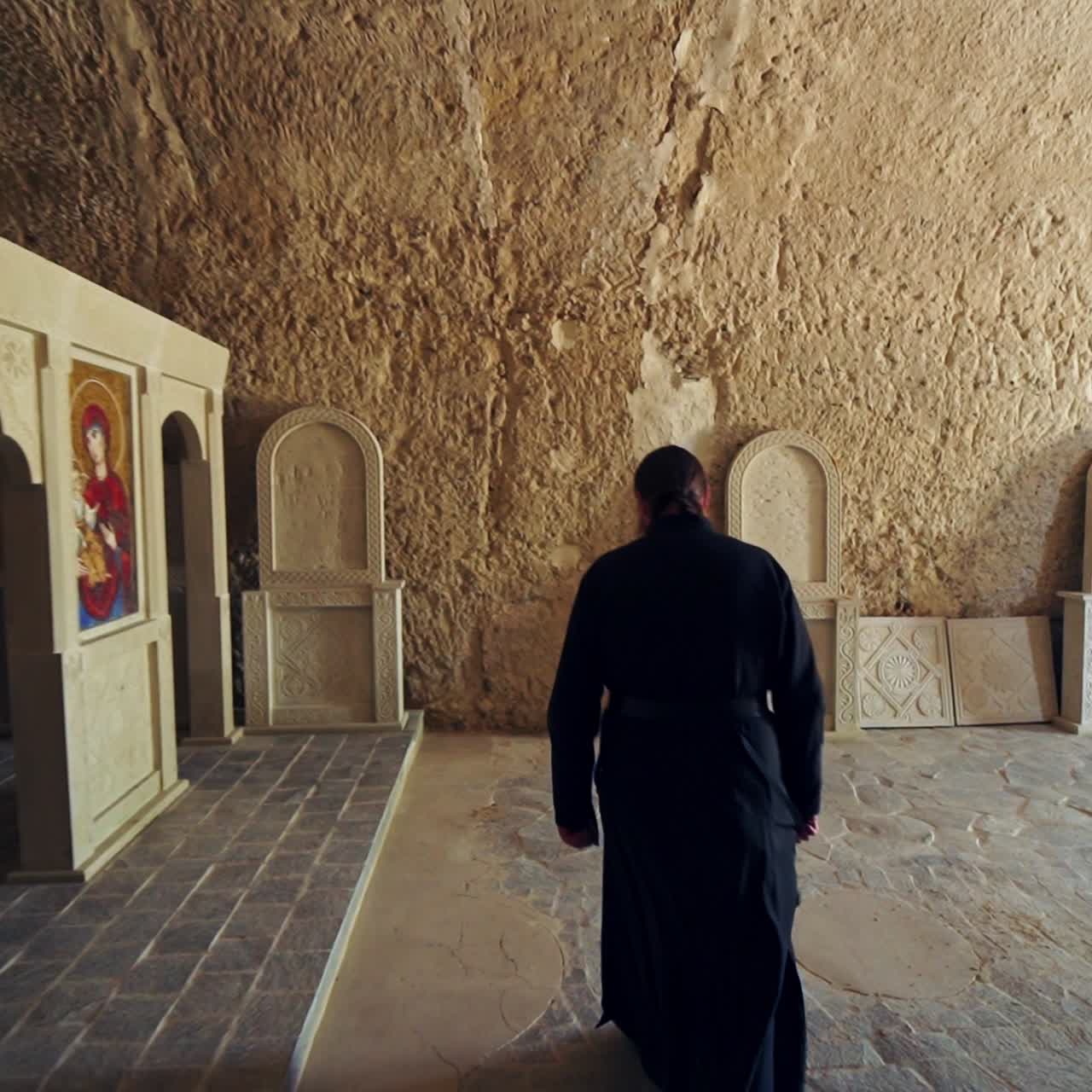 Male silhouette moving by the long corridor with stone walls. Rear view of the monk in black robe entering the chapel in rock monastery