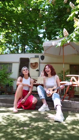 two women playing ukulele in a park outside a camping trailer