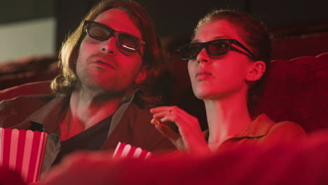 vista de cerca de una pareja con gafas 3d sentada en el cine mientras ven una película y comen palomitas de maíz con las luces apagadas