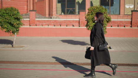 elegant lady adjusts black handbag strap while walking past red brick building with decorative plant and stylish window displays, wearing black coat and boots on a bright urban street in daylight