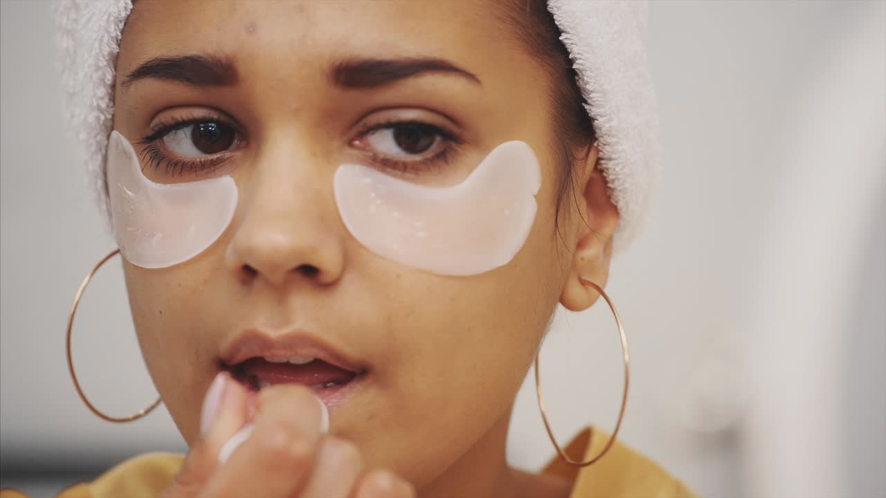 Closeup portrait of beautiful woman after bath with towel on head puts patches under the eyes from wrinkles and dark circles. Attractive female in bathrobe makes beauty treatments on the face at home.