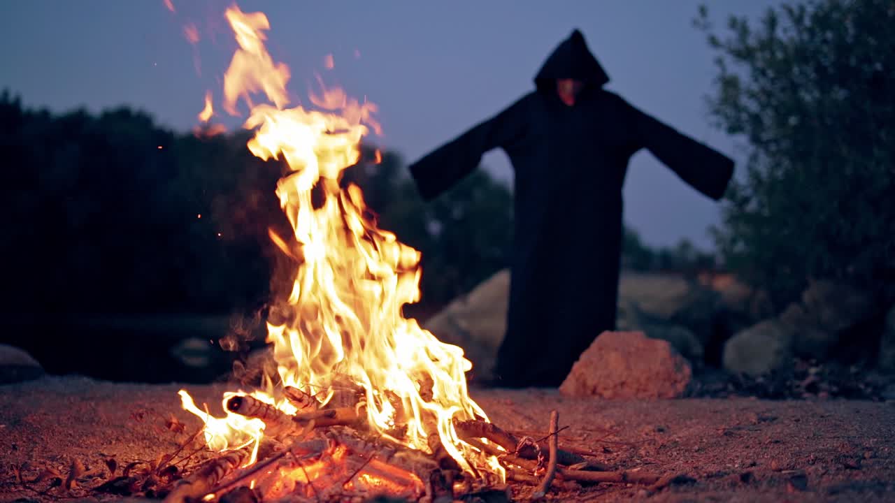 Monk pray at the campfire. Man in a cassock performs around fire