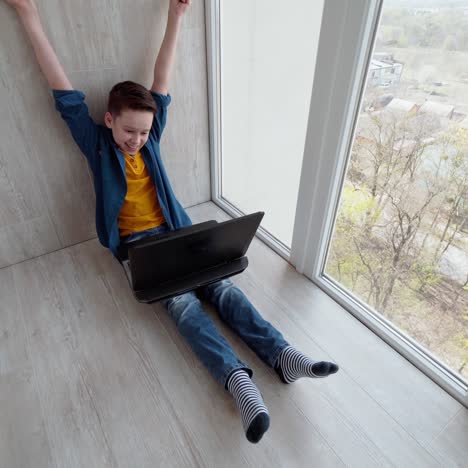 teenager with a laptop on a balcony. boy sitting on the floor near the window and playing video games. happy teen wins the game. top view.