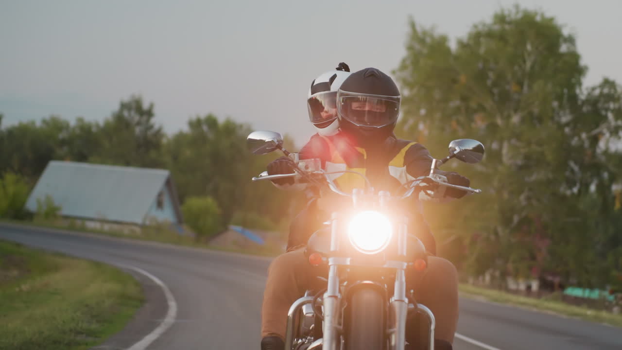 Biker wearing black helmet and leather jacket with passenger in white helmet makes bend on uphill countryside road, passing houses and trees in evening light