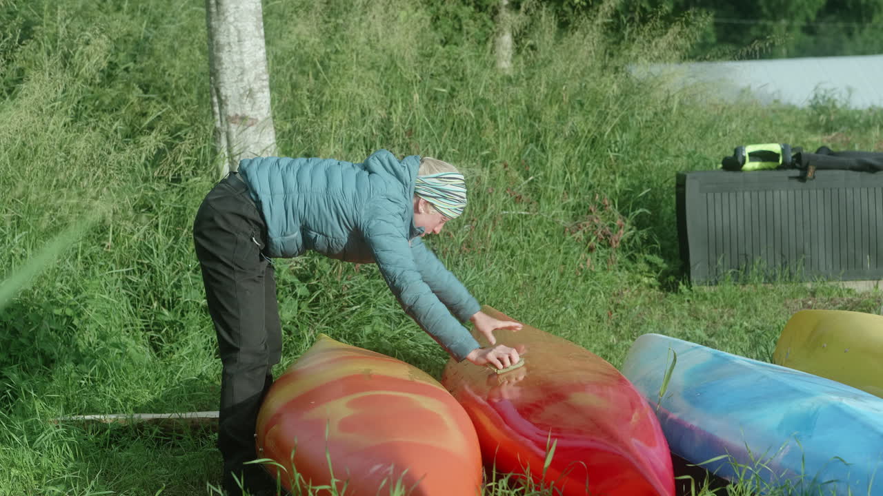 Nordisk blond jente rengjør kajakkbåter som ligger på bakken, vakker solrik morgenstemning, Finland, Kvarken skjærgård
