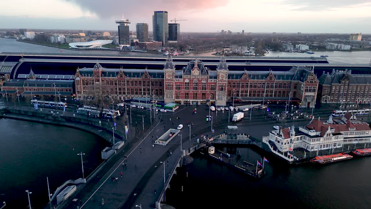 Visitors admire the beautiful architecture of Amsterdam's central station while enjoying the waterfront views at dusk. The vibrant city life unfolds as boats float nearby
