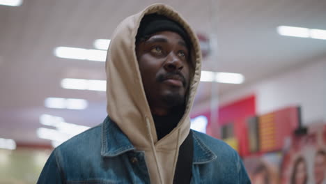 close shot of male walking through bright indoor mall wearing beige hoodie and denim jacket with beanie under hood and backpack strap visible side profile in motion against blurred store lights