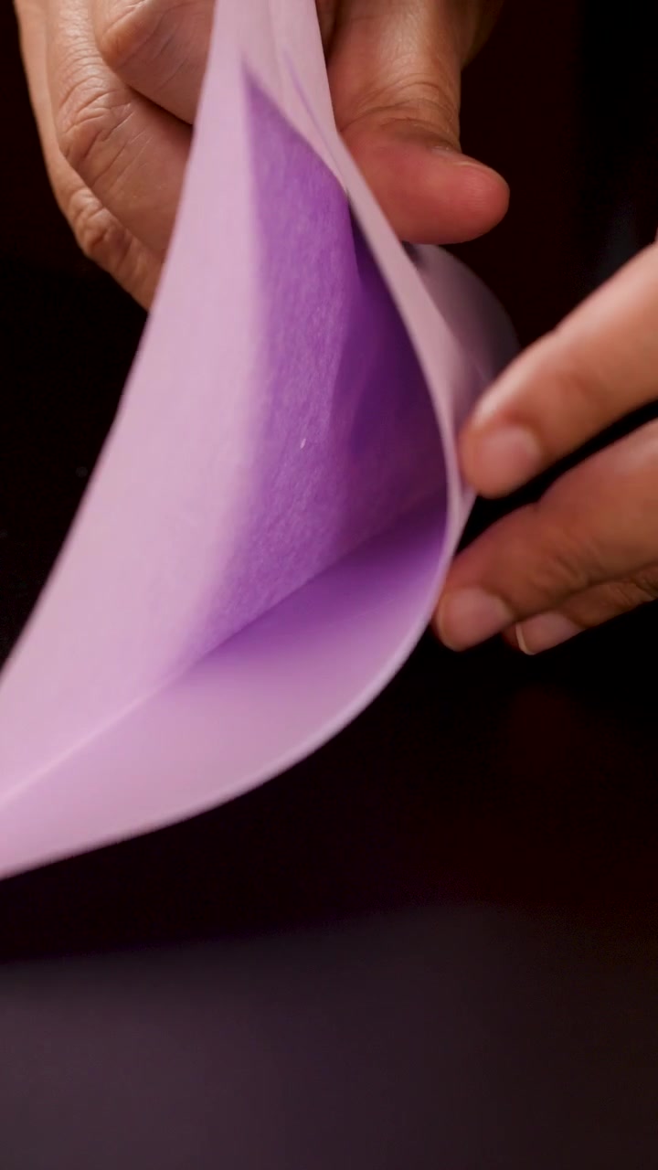 Voksne hænder folder forsigtigt hvidt papir til en origamibåd på en mørk bordplade.