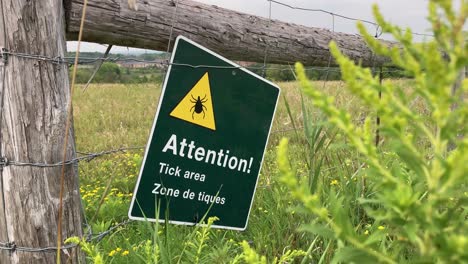 tick area warning sign on rural park trail fence with tall grass in farmland field