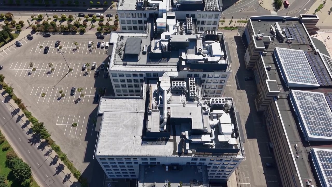Filmagem aérea dinâmica captura edifícios de escritórios elegantes com painéis solares e um estacionamento vazio sob luz solar intensa. Vídeo de drone de alta resolução, sombras nítidas, arquitetura geométrica