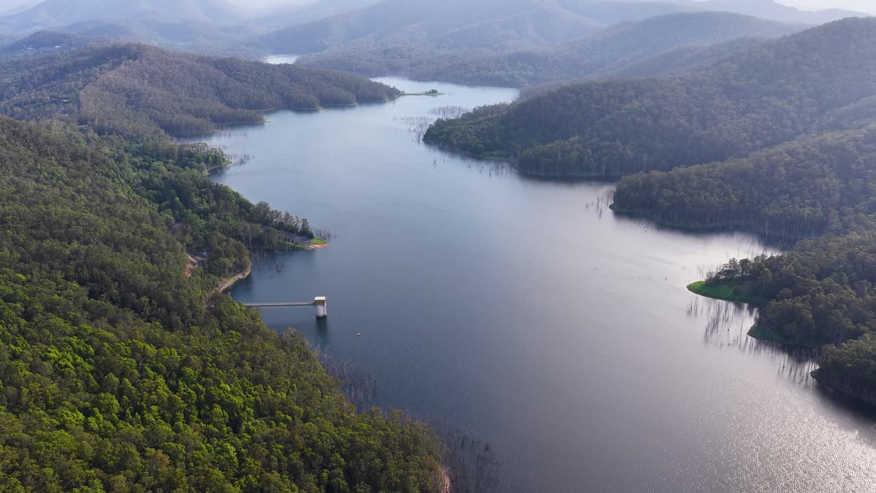 Drone glides over tranquil mountain reservoir, lush forest, and rolling hills in soft daylight