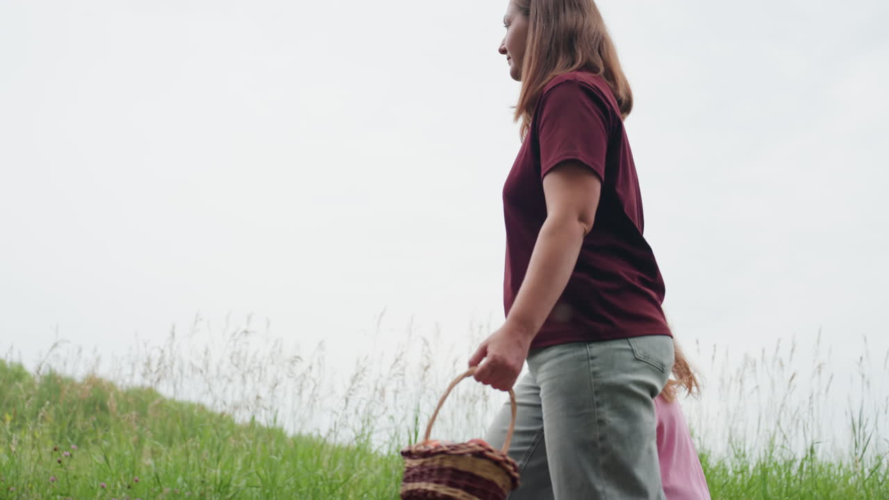 madre bianca che trasporta un cesto di vimini attraverso il prato, primo piano della mano che tiene il cesto di vimini, passo deciso tra l'erba alta, bambino al seguito, atmosfera da picnic rustico, toni naturali e gita in famiglia spontanea