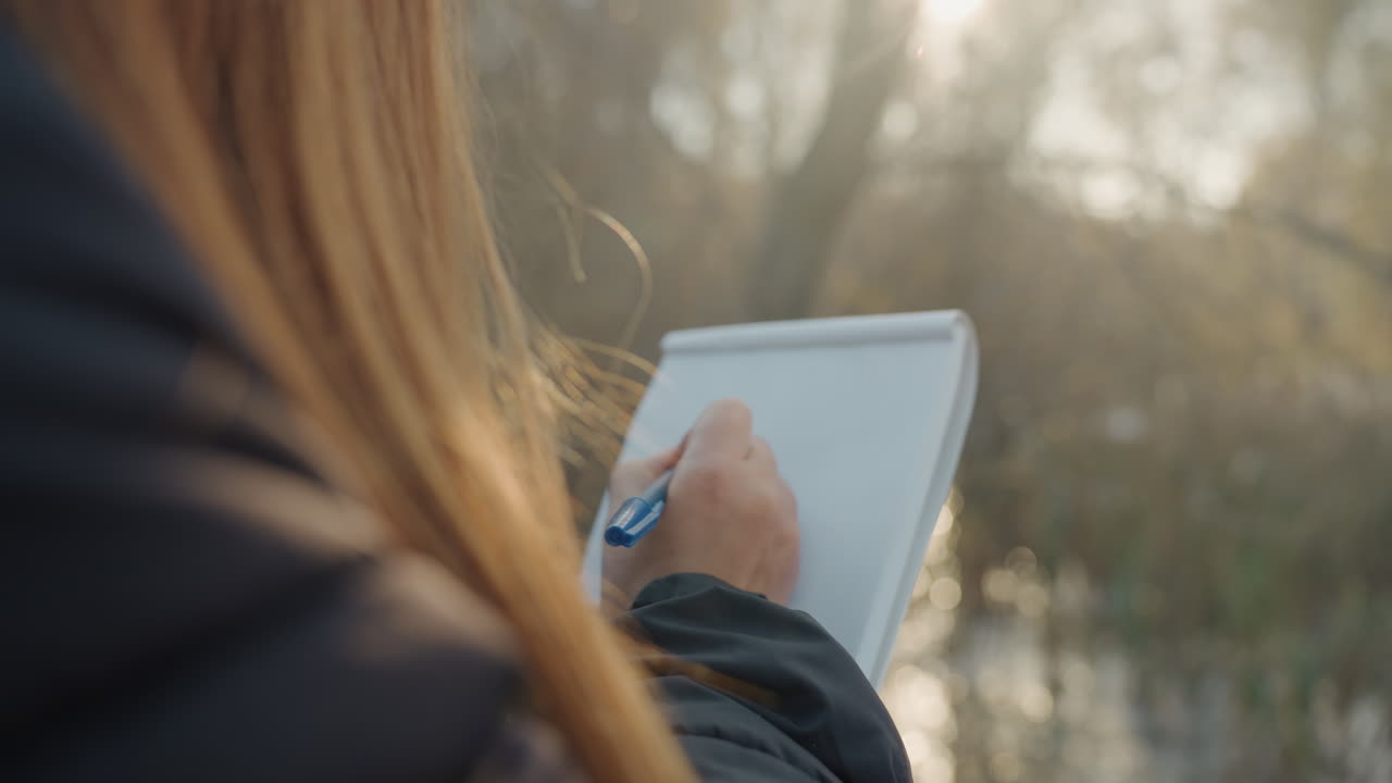 Lady captures notes beside flowing water. Caucasian woman jotting quick sketches in peaceful outdoor setting. Woman with long hair sketches and notes beside river during tranquil golden sunset