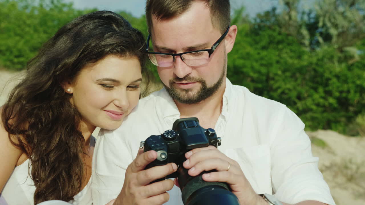 um jovem casal olha as fotos em uma câmera digital senta-se ao lado da praia ótimas lembranças de h
