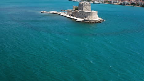 Volando-Sobre-El-Mar-Azul-En-Calma-Hacia-El-Castillo-De-Bourtzi-En-Un-Día-Soleado-En-Nafplio,-Grecia